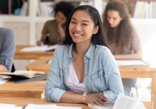 Smiling smart female asian teen girl looking at camera sitting at desk studying in classroom with diverse classmates group, happy chinese college university student in international class portrait