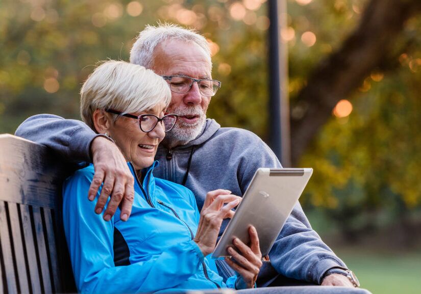 Smiling senior active couple sitting on the bench looking at tab